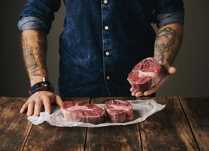 Brutal butcher with tattooed hands offers a piece of great raw meat steak on camera, other steaks in white craft paper on vintage old wooden grunge table. unrecognizable