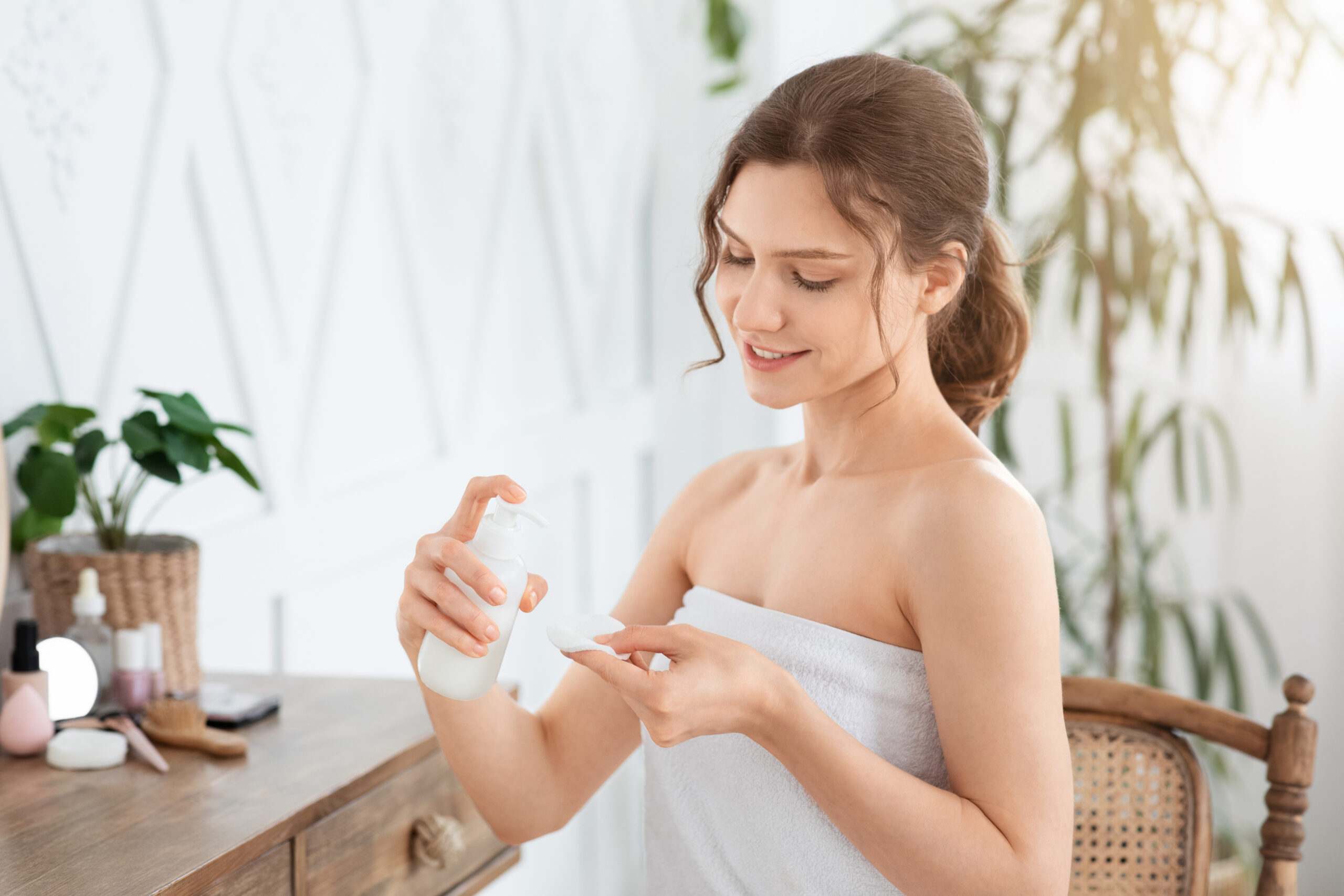 Side view of cheerful woman cleaning her face, using cotton pads and cleansing product, looking at mirror in bedroom. Young attractive lady using face toner and cotton pad, home interior, empty space